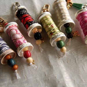May include: A selection of handmade keyrings, each with a miniature thread spool. The spools are wrapped in patterned fabric and decorated with wooden beads and a gold-tone clasp. The spools have a measurement of 250 m - 274 yards.