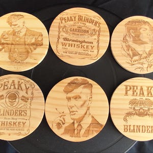 Może przedstawiać: Zestaw sześciu drewnianych podkładek z grawerowanymi wzorami. Podkładki przedstawiają portrety i tekst związane z "Peaky Blinders" i "Whiskey". Podkładki są okrągłe i wykonane z jasnego drewna.