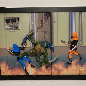 May include: A framed diorama featuring four action figures from the Teenage Mutant Ninja Turtles franchise. The figures are posed in a fighting stance, with one figure holding a gun and another holding a sword. The diorama is set against a backdrop of a city street.