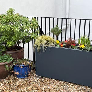 May include: A garden scene with a large, dark grey rectangular planter box filled with colourful flowers and greenery. Several terracotta and blue ceramic pots with plants are also visible. The ground is covered in small pebbles, and a black railing is in the background.