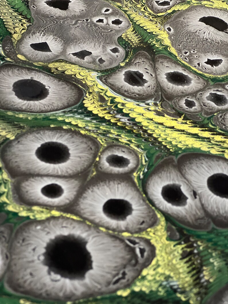 May include: Close-up view of fluid acrylic pour painting. The artwork features a mesmerizing pattern of dark gray and black circular shapes resembling eyes, set against a backdrop of swirling green and gold. The gray shapes have a textured, almost organic appearance, with subtle variations in tone and shading. The green and gold background creates a dynamic contrast, enhancing the overall visual impact of the piece. The colors are rich and vibrant, and the texture is smooth yet dynamic, giving the painting a three-dimensional quality. The image showcases the unique characteristics of fluid acrylic pouring, highlighting the unpredictable yet beautiful results of this technique.