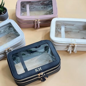 May include: Four zippered cosmetic bags in various colors: light pink, light gray, white, and navy blue. Each bag has a clear top, gold-colored zipper, and a personalized monogram. The bags are rectangular and appear to be made of leather or a similar material.