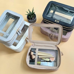 May include: Three clear makeup bags with gold zippers. The bags are in different colors: light gray, light pink, and black. The black bag has the initials KH on it. The light pink bag has the words "JAZD Beauty" on it. The light gray bag has a clear window on the front. The bags are all open and show the inside of the bags. The light pink bag has makeup brushes and other makeup items inside.