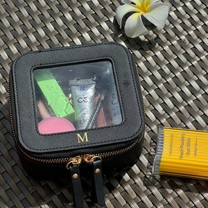 May include: Black square makeup bag with a clear window showing various cosmetics, including a green tube, a pink tube, a black tube, and a pink sponge. The bag has a gold zipper and the letter "M" is printed on the front.