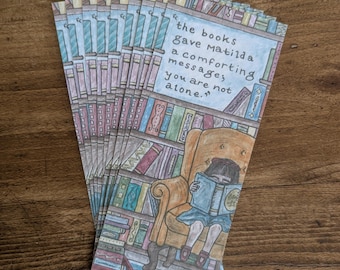 May include: A stack of bookmarks featuring a girl sitting in a chair reading a book in a library. The text on the bookmark reads "The books gave Matilda a comforting message; you are not alone."
