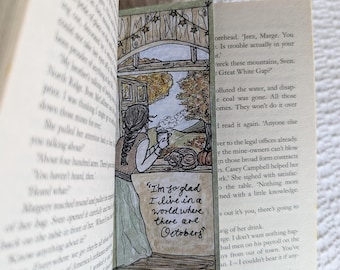 May include: A book with a hand-drawn bookmark featuring a woman sitting in a window with a cup of tea. The bookmark has the text "I'm so glad I live in a world where there are Octobers."