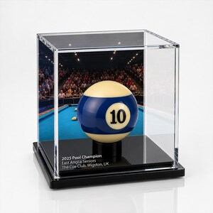 Op de afbeelding: Een nummer 10 biljartbal in een doorzichtige acryl kubus op een zwarte voet. De behuizing heeft een geprinte achtergrond van een biljartzaal en de tekst 2025 Pool Champion, East Anglia Seniors, The Cue Club, Wigston, UK.