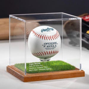 May include: A baseball in a clear display case. The white ball has red stitching and the Rawlings logo. The case sits on a wooden base with a patch of artificial turf. Text on the base reads "Logan Mitchell Walk-Off Home Run Falcons vs. Eastside Titans May 3, 2025 - Riverside Stadium."