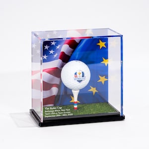 Pode incluir: Uma bola de golfe num tee dentro de uma vitrine transparente. O fundo apresenta as bandeiras americana e da União Europeia. O texto na base diz "The Ryder Cup, Bethpage Black, New York, Team USA vs Team Europe, September 26th-28th, 2025."
