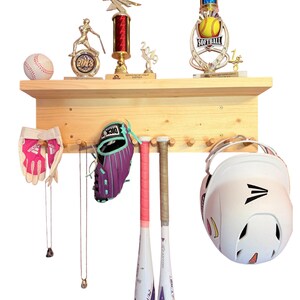 May include: A wooden wall shelf with hooks for hanging sports equipment. The shelf has two baseball bats, a baseball glove, a baseball helmet, and a baseball. There are also several trophies on the shelf.