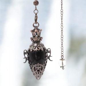 Könnte beinhalten: Ein schwarzes Pendel mit aufwendiger Metallarbeit. Das Pendel hat eine Tropfenform mit spitzem Ende und dekorativer Kappe. Eine Kette mit einer kleinen Kristallperle ist befestigt. Das Metall hat eine Bronze-Optik, das Design beinhaltet Sterne und geometrische Formen.