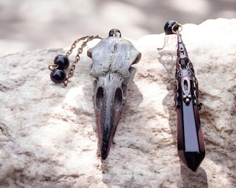 May include: Two pendants, one with a detailed, gray, bird skull and the other with a black, faceted crystal, both with ornate metal accents.