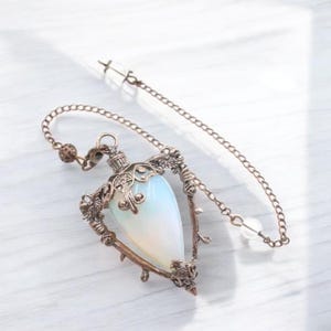 May include: A pendulum with a teardrop-shaped, opalescent crystal encased in an ornate, bronze-colored metal frame. The frame features decorative floral and vine-like details. A chain with a small crystal bead and a larger clear crystal at the end is attached.
