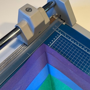 Pode incluir: Um close-up de um cortador de papel com uma lâmina prateada e um tapete de corte azul. O cortador está sendo usado para cortar uma pilha de papel colorido, incluindo roxo, azul e verde.
