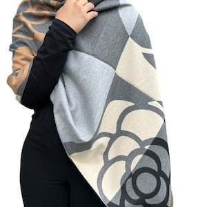 May include: A patterned shawl in shades of gray, beige, and black. The shawl features a large floral design and geometric shapes. The person is wearing a black long-sleeved shirt. The shawl is draped over the shoulders.