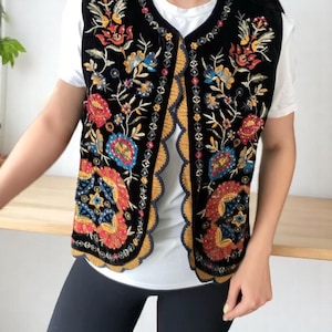 Può includere: Un gilet in velluto nero caratterizzato da intricati ricami floreali multicolori con paillettes. Il capo presenta un bordo anteriore smerlato rifinito in giallo senape e blu scuro, indossato sopra una t-shirt bianca e leggings neri.