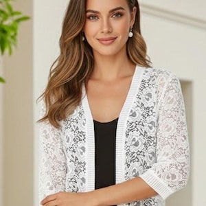 May include: A white open-front cardigan featuring a delicate floral lace pattern throughout the body and sleeves. The garment has three-quarter length sleeves and a ribbed knit trim along the front opening, cuffs, and hemline, worn over a black top.