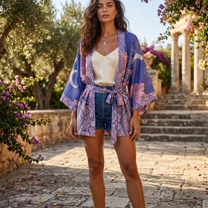 May include: A blue and purple kimono-style jacket featuring a celestial print with moons and sunbursts, paired with a white camisole and blue denim shorts. The jacket has wide sleeves, a matching tie belt at the waist, and a floral border pattern.