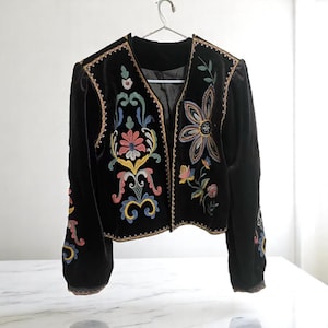 May include: Black velvet jacket with colourful floral embroidery. The jacket has a V-neck, long sleeves, and intricate embroidery in shades of pink, blue, yellow, and green. The jacket is hanging on a metal hanger.