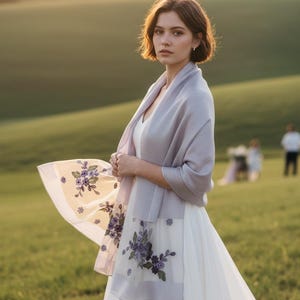 Puede incluir: Un chal de color lavanda claro sobre un vestido blanco. El chal presenta paneles transparentes en los extremos decorados con un intrincado bordado floral morado, que incluye pequeñas hojas verdes y pétalos de color morado oscuro.