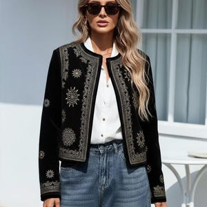 May include: Black velvet jacket with gold embroidery featuring sunburst and floral designs. The jacket has a round collar and is worn over a white shirt and blue jeans. The jacket's design is on the front and sleeves.