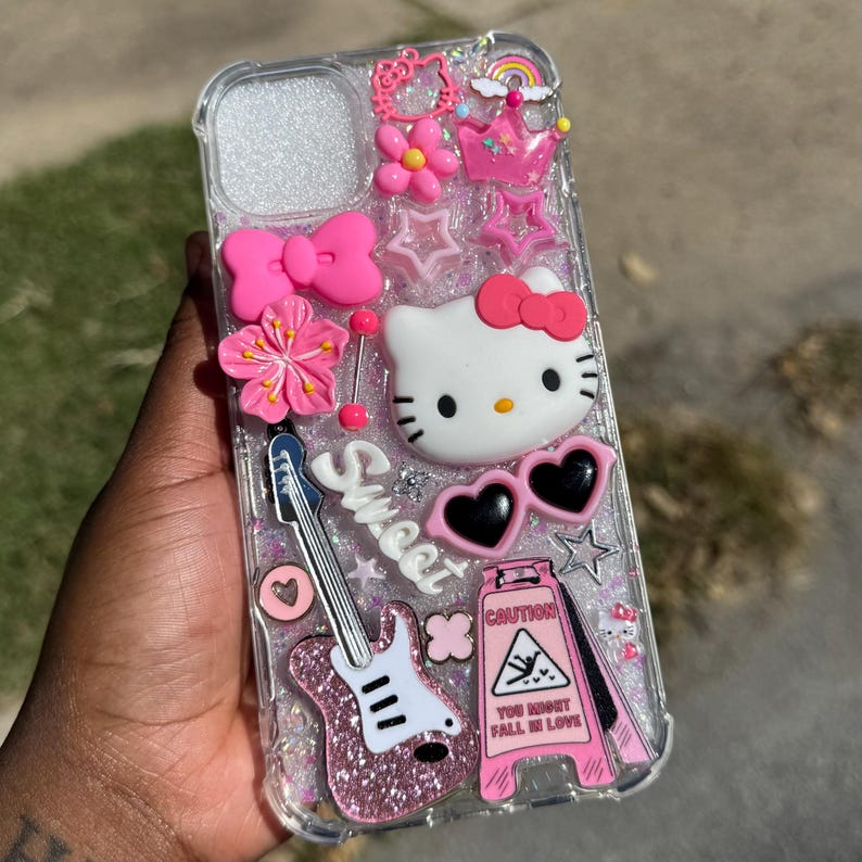 Pode incluir: Capa de telefone transparente decorada com enfeites rosa e brancos. Apresenta um rosto de Hello Kitty, &oacute;culos de sol em forma de cora&ccedil;&atilde;o, uma guitarra e a palavra "Sweet". Inclui um sinal de aviso que diz "You Might Fall In Love".