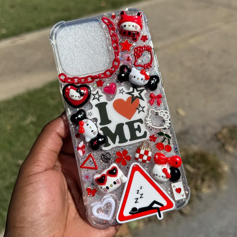 May include: Clear phone case decorated with various charms, including Hello Kitty figures, hearts, stars, and a road sign. The case has the words "I ME" in the center, with red, black, and white accents. The case is held in a hand.