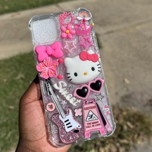 Pode incluir: Capa de telefone transparente decorada com Hello Kitty rosa, flores, la&ccedil;os, estrelas e uma guitarra. A capa tamb&eacute;m apresenta um sinal de aviso que diz "You Might Fall in Love" e a palavra "Sweet".