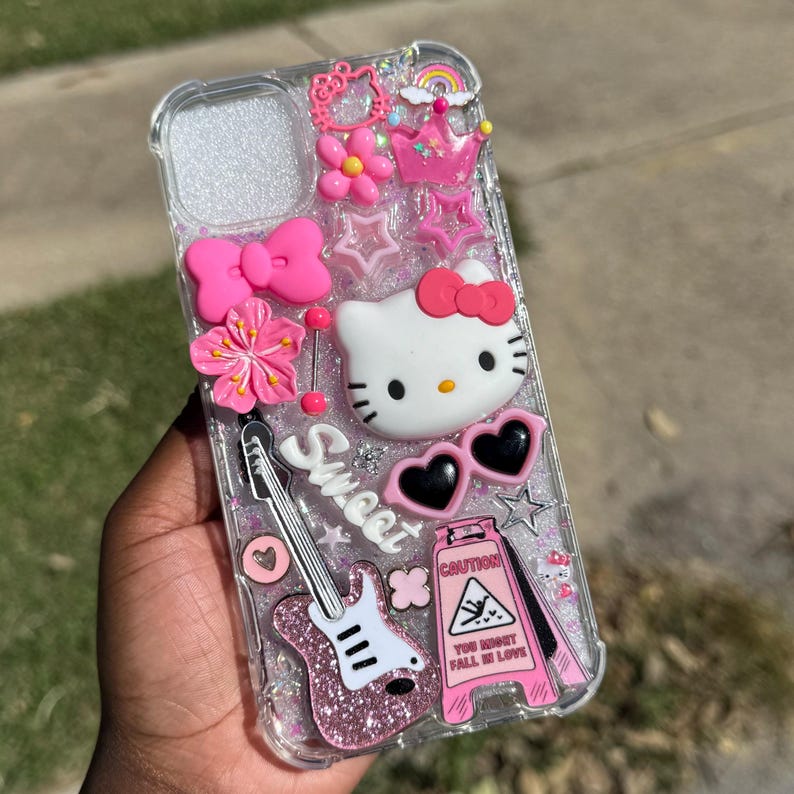 Pode incluir: Capa de telefone transparente decorada com Hello Kitty rosa, flores, la&ccedil;os, estrelas e uma guitarra. A capa tamb&eacute;m apresenta um sinal de aviso que diz "You Might Fall in Love" e a palavra "Sweet".