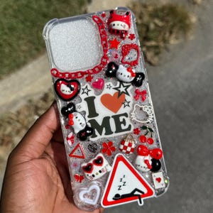 Puede incluir: Funda de teléfono transparente decorada con varios abalorios, incluyendo figuras de Hello Kitty, corazones, estrellas y cerezas. La funda presenta el texto "I ❤️ ME" y un diseño de cadena roja. La funda está llena de purpurina.