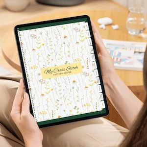 May include: A digital tablet displaying a cross stitch pattern keeper app with a white background featuring a watercolor floral print of daisies and wildflowers. A yellow rectangular label in the center reads My Cross Stitch PATTERN KEEPER.
