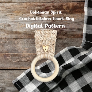 Peut inclure: Un anneau de serviette de cuisine en crochet beige et blanc avec un petit charme en forme de cœur doré. L'anneau est fait avec un anneau en bois et est posé sur un tissu à carreaux noir et blanc.