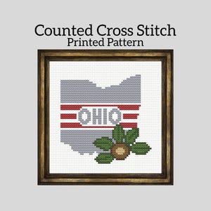 May include: A framed counted cross stitch pattern featuring the outline of Ohio in gray, with red and white stripes and the word "OHIO". A green and brown floral design is at the bottom. The text "Counted Cross Stitch Printed Pattern" is at the top.