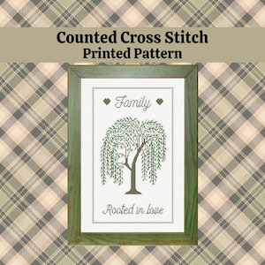 May include: A framed counted cross stitch pattern featuring a weeping willow tree. The text includes "Family" and "Rooted in love." The frame is green, and the background is a plaid pattern in shades of green, tan, and gray.
