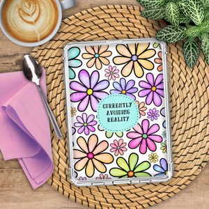 May include: A clear tablet case featuring a repeating pattern of multi-colored flowers in shades of pink, purple, yellow, orange, and green. In the center, a light blue circular emblem displays the black text: CURRENTLY AVOIDING REALITY.