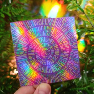 May include: Square holographic sticker with a tie-dye spiral design in shades of purple, pink, and teal. The sticker reflects rainbow colors. The design features a series of concentric circles with a dotted pattern.