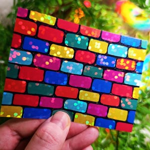 May include: A rectangular card with a colorful brick pattern. The bricks are various colors, including red, blue, green, yellow, and pink, with a shiny, iridescent finish. The card is held in a hand.