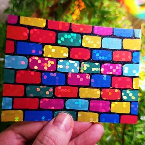 May include: A rectangular card with a colorful brick pattern. The bricks are various colors including red, yellow, blue, green, and pink, with a black outline. The bricks have a glittery, reflective surface.