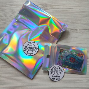 May include: Iridescent holographic bags with a small clear bag containing a colorful card. Two circular stickers feature a cat in a UFO with the text "ufo cat designs" and "freebies for you".