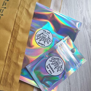 May include: Two iridescent cellophane bags with a circular sticker that says "ufo cat designs" and "freebies for you". The bags are inside a brown paper envelope. The background is a light wood grain.