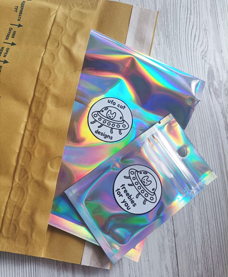 May include: Two iridescent cellophane bags with a circular sticker that says "ufo cat designs" and "freebies for you". The bags are inside a brown paper envelope. The background is a light wood grain.