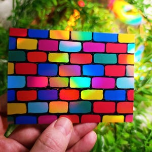 May include: A rectangular card featuring a vibrant brick wall design. The bricks are a mix of bright colors including red, yellow, blue, and pink, outlined in black. The card is held against a blurred green background.