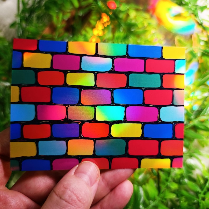 May include: A rectangular card featuring a vibrant brick wall design. The bricks are a mix of bright colors including red, yellow, blue, and pink, outlined in black. The card is held against a blurred green background.