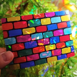 May include: A rectangular card with a colorful brick pattern. The bricks are various colors including red, blue, yellow, and green. The surface of the bricks has a holographic, iridescent effect, creating a shimmering appearance. The card is held in a hand.