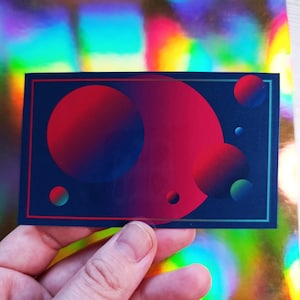 May include: A rectangular sticker featuring a design of overlapping circles in shades of red, blue, and purple against a dark blue background. The sticker has a thin teal border and is held in a hand.