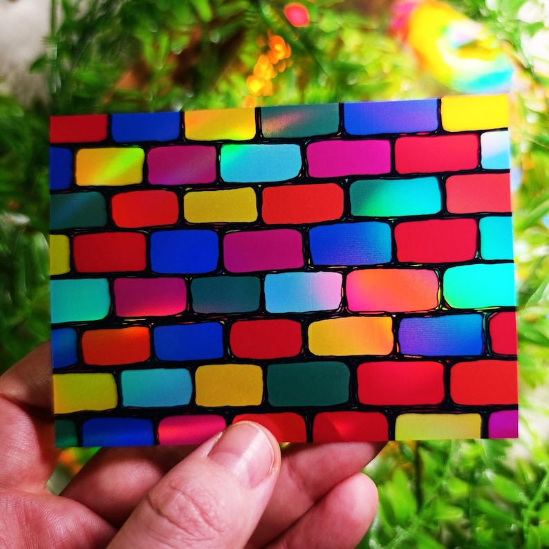 May include: A rectangular card featuring a vibrant, colorful brick wall design. The bricks are a mix of bright hues including red, yellow, blue, and pink, with black outlines. The card is held in a person's hand, with a blurred green background.