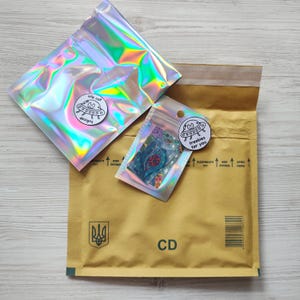 May include: Two iridescent cellophane bags and a small clear bag with a sticker on a brown envelope. The bags have stickers with a cartoon UFO and the words "ufo cat designs" and "freebies for you". The envelope has the letters "CD".