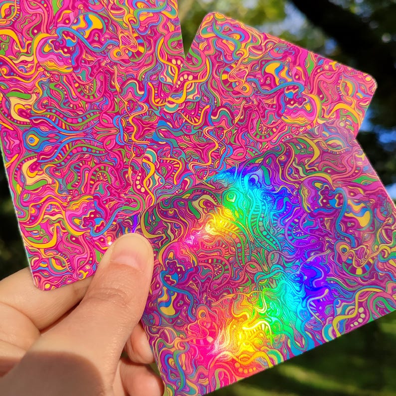 May include: Two square cards with a vibrant, psychedelic design. The cards feature a swirling pattern of pink, yellow, blue, and green, with a holographic rainbow effect. The cards are held up against a blurred green background.