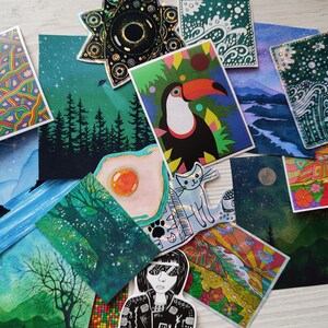 May include: A collection of colorful art prints and stickers. The prints feature various illustrations, including a toucan, landscapes, and abstract patterns. A star-shaped sticker and a cat sticker are also visible.