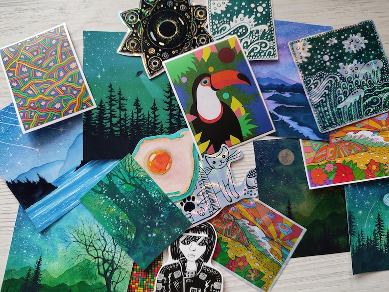 May include: A collection of colorful art prints and stickers. The prints feature various illustrations, including a toucan, landscapes, and abstract patterns. A star-shaped sticker and a cat sticker are also visible.
